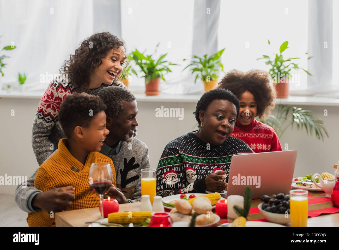 middle aged african american woman talking during video call near cheerful family and served dinner Stock Photo
