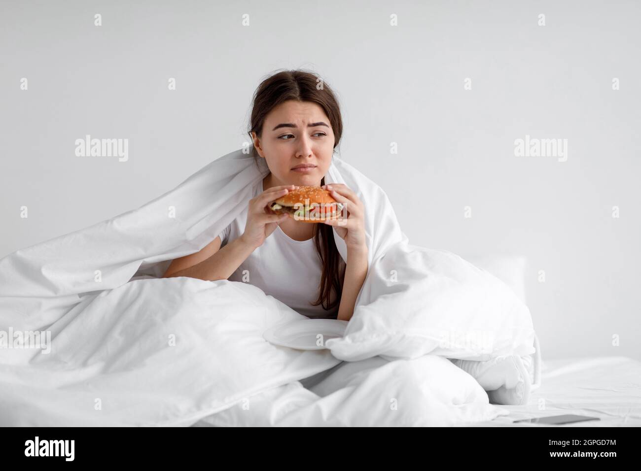 Sad upset depressed european millennial woman eating burger in bed under duvet at home alone Stock Photo