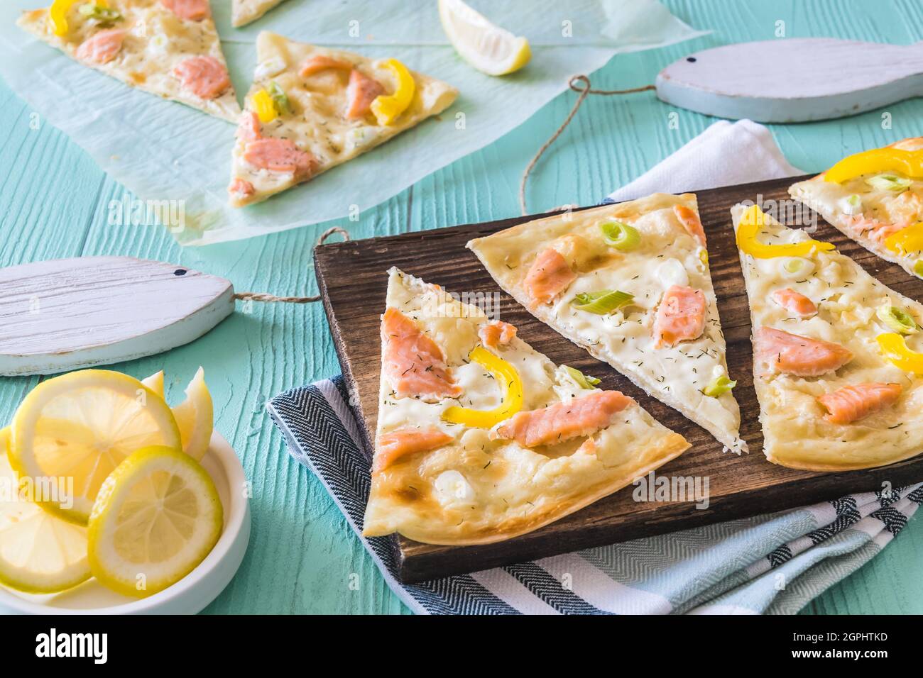 Pieces of tarte flambee with salmon, yellow pepper and spring onions on a wooden board on light green background Stock Photo