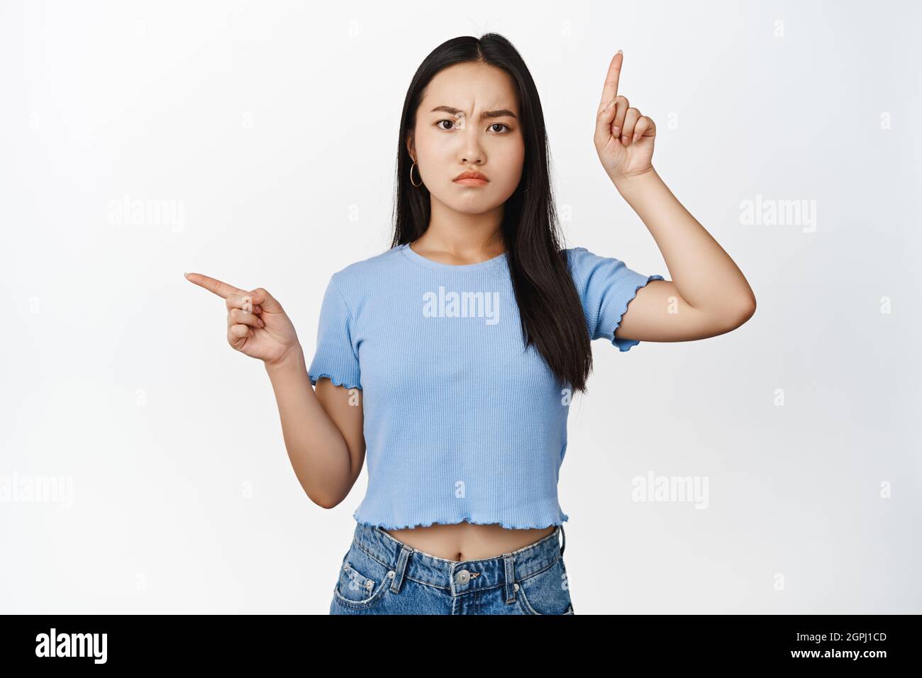 Angry teen asian girl pointing sideways, sulking and frowning upset, complaining at something, standing in blue t-shirt over white background Stock Photo