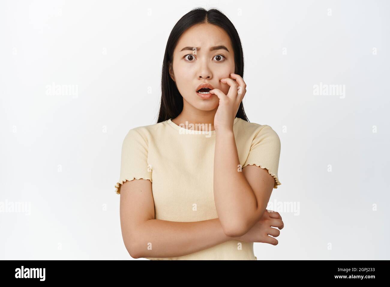Anxious asian girl biting fingernails and looking worried, being in trouble, panicking, standing over white background Stock Photo