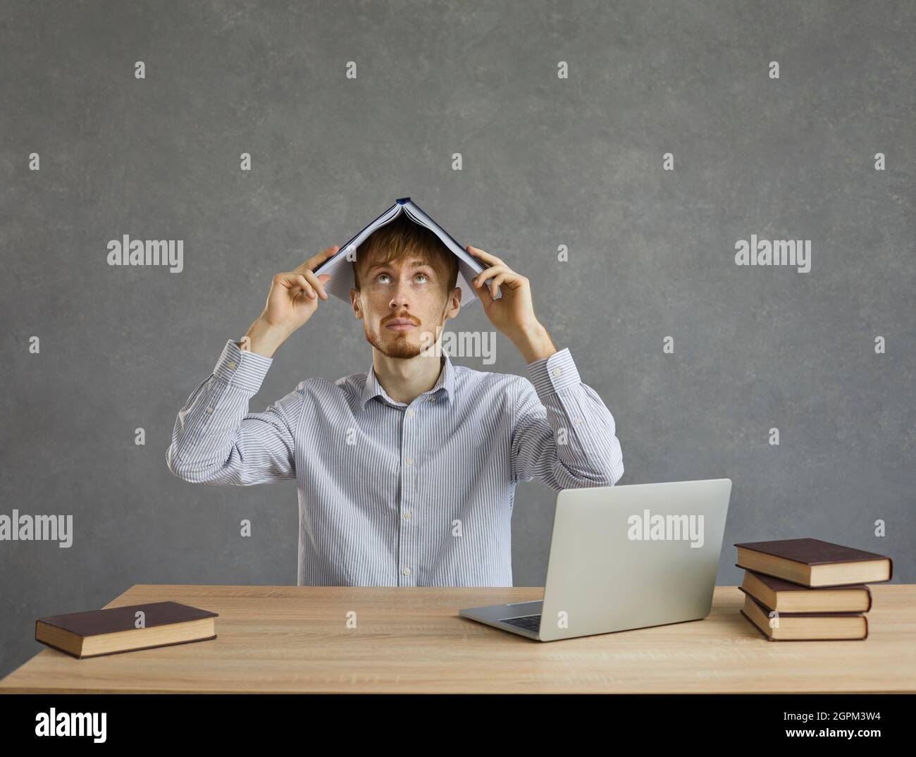 Male student with a pleading look on his face looking up holding a book on his head. Stock Photo