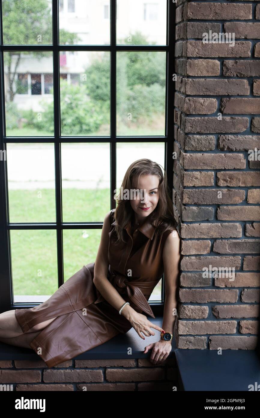 Beautiful young woman in brown leather dress sits on the windowsill ...