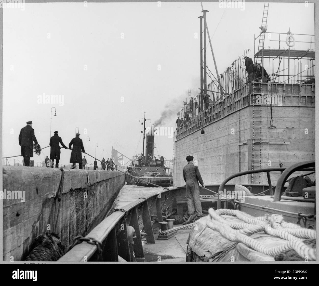 A view from a boat showing the cooling water intake caisson for Coryton Oil Refinery leaving Tilbury Docks, with people on the side of the lock using ropes to guide the structure. During the Second World War, John Laing &amp; Son Ltd had built some of the floating caissons for the Mulberry harbour which were used in the D-day landings. This 4,200 ton concrete water intake caisson was built along the lines of the Mulberry harbour and was towed 8 miles down the Thames from Tilbury Dock to be installed as part of Coryton Oil Refinery in March 1952. It was built between 1951 and 1952. Stock Photo