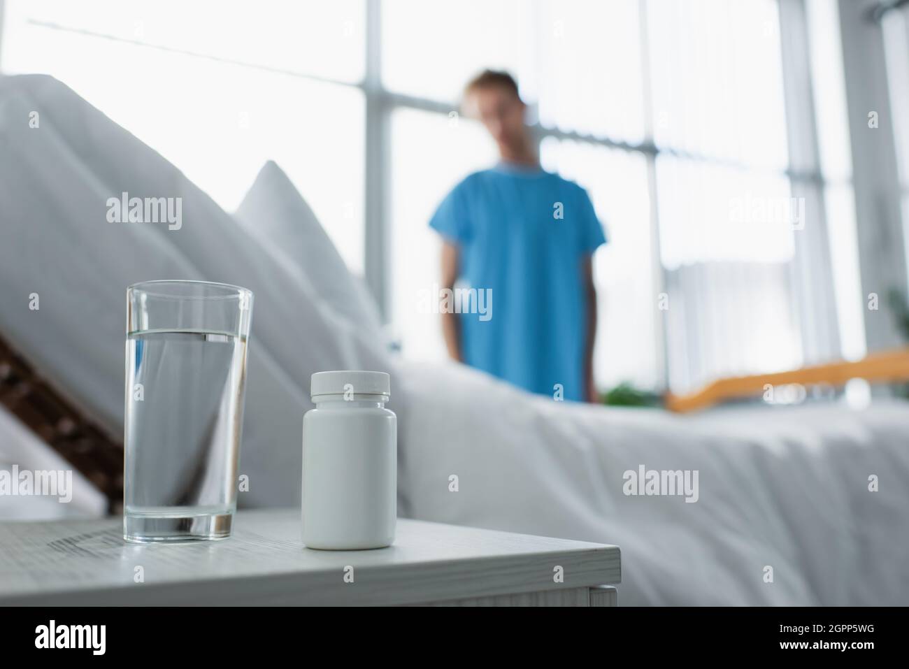 bottle with medication and glass of water on bedside table near blurred patient in hospital Stock Photo
