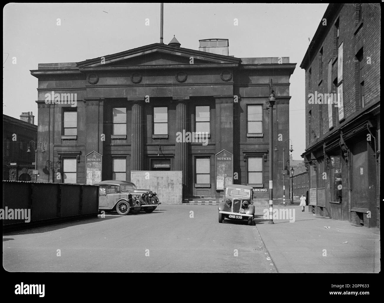 Town Hall, Bexley Square, Salford, 1942. An exterior view of the Town Hall, showing the south facade with two contemporary cars in the foreground. The hall was built by Richard Lane between 1825 and 1827, and the south facade has two storeys and five bays, with a three bay Doric portico with giant columns, a pediment and frieze with four relief wreaths. In the image the windows have blackout fabric covering them, and a temporary brick wall over the main entrance. The street furniture is painted with black and white stripes, to faciliate night-time driving during World War II. Stock Photo