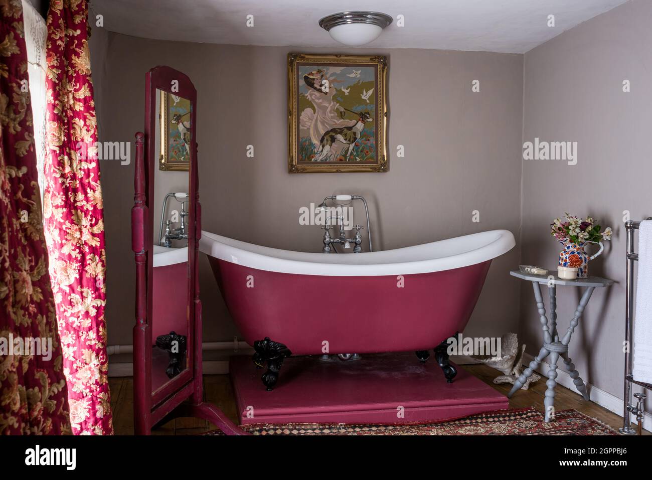 Bold pink bath with vintage curtains and needlework panel in Suffolk cottage. Stock Photo