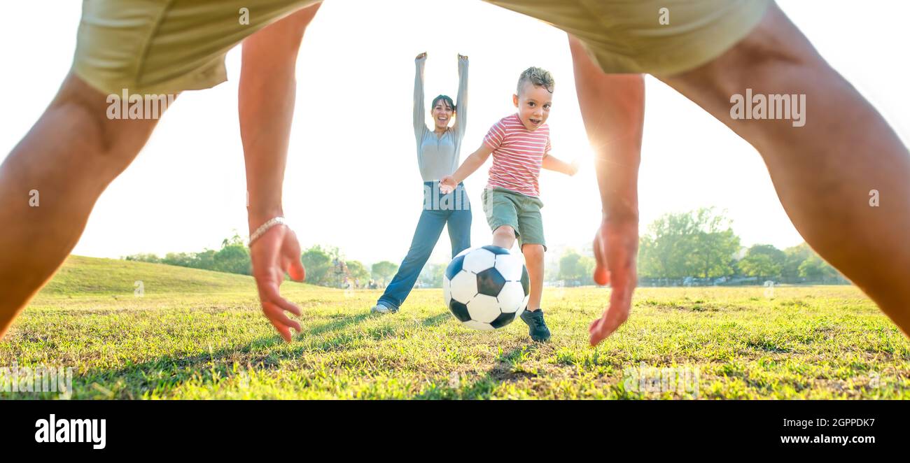 young happy family playing football outdoors into a public park garden. mom, dad and little child having fun together. cute kid kicking a ball Stock Photo