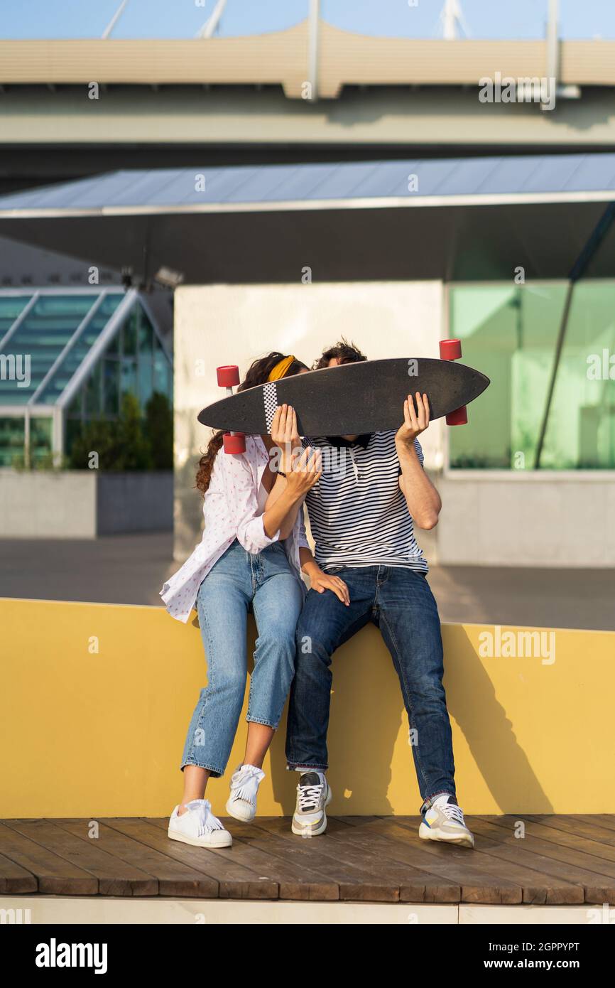 Joyful couple kiss hiding above longboard. Playful man and woman in love together hold skateboard Stock Photo