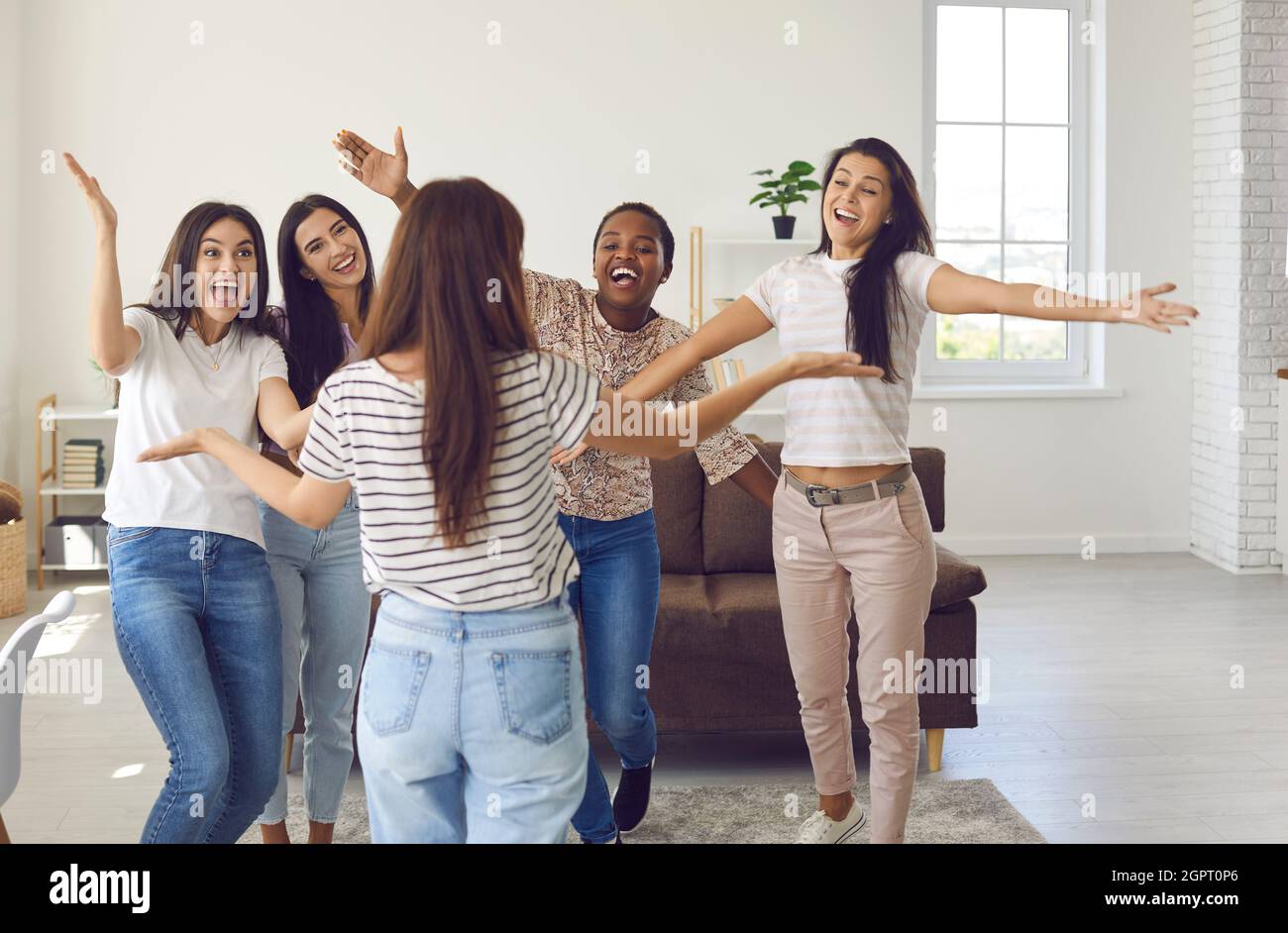 Happy group of young multinational women greet and hug their female friend during meeting at home. Stock Photo