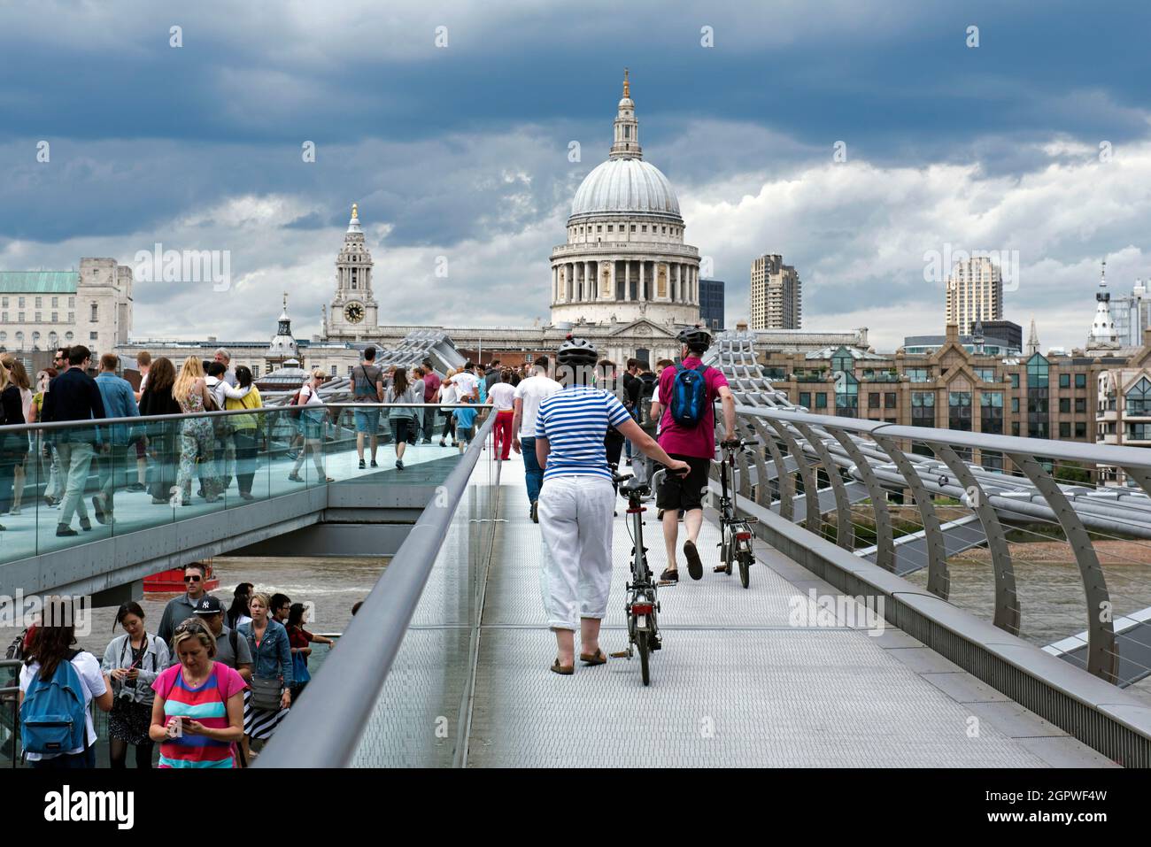 People crossing London Millennium Footbridge or Bridge across the Thames in summer some pushing bikes with St. Paul's Cathedral in the background. Stock Photo