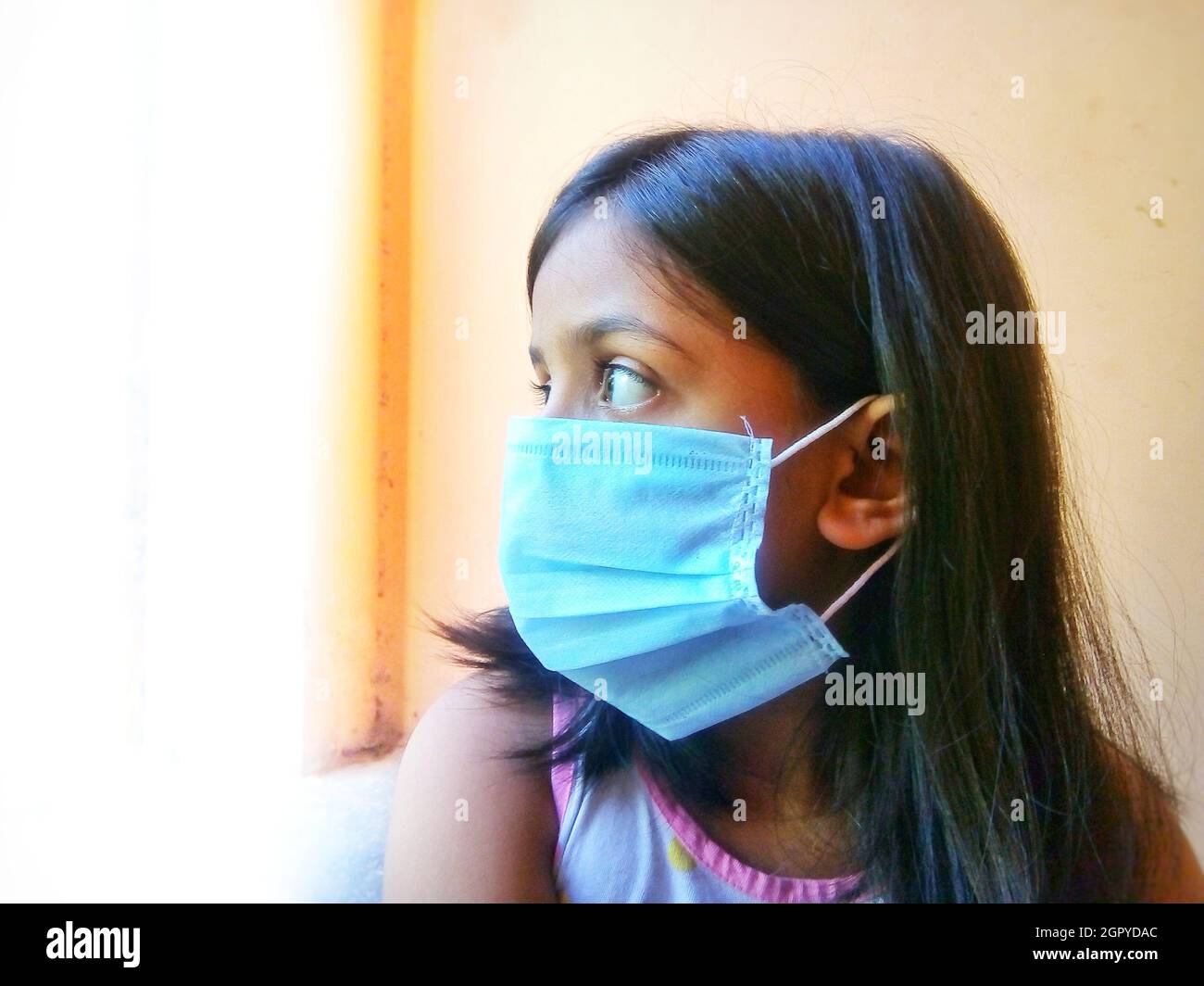 Portrait Of A Girl Wearing Protective Face Mask While Looking Outside The Window, India Stock Photo