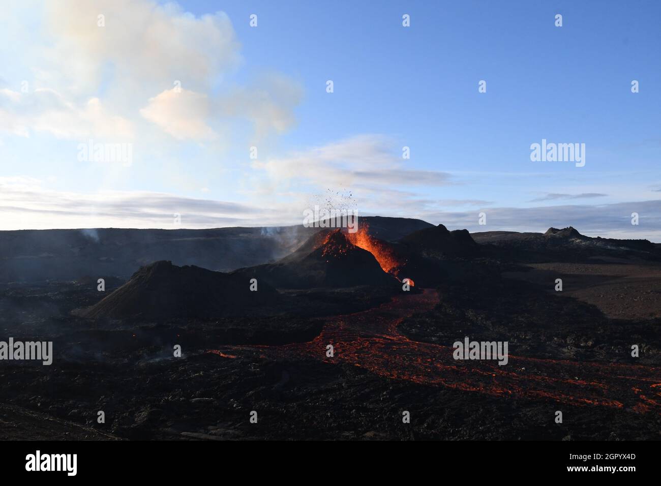 The lava field at Fagradalsfjall, Iceland. The active vent has orange molten lava erupting from it and white volcanic gas rising. Black lava field. Stock Photo