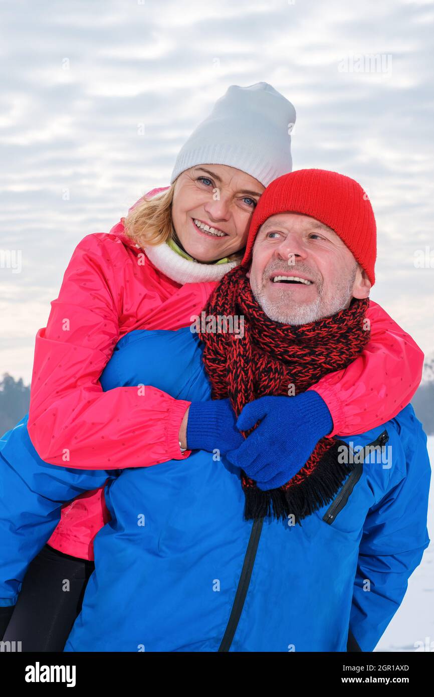 Smiling senior man giving mature woman piggyback in snowy winter park ...
