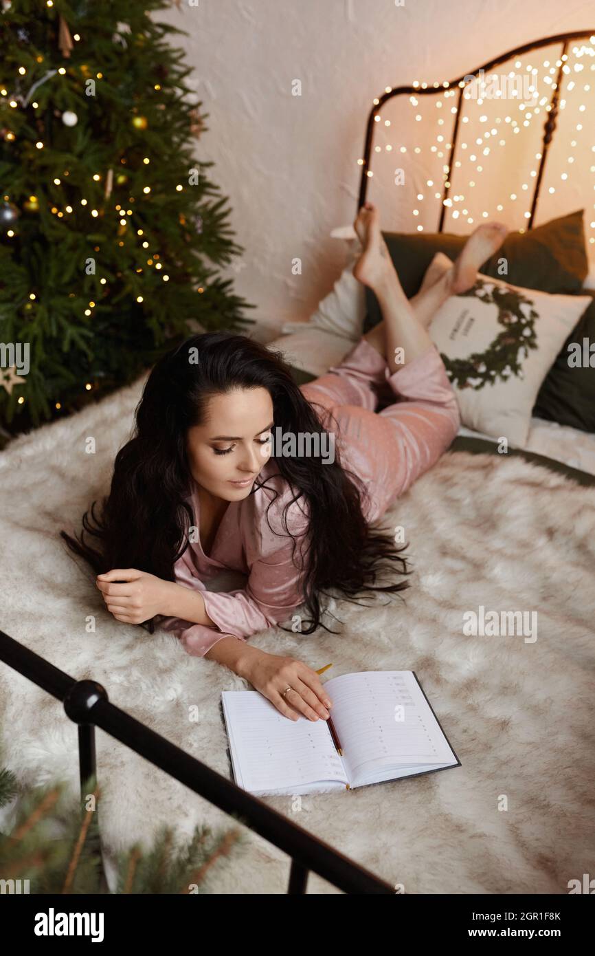Pretty brunette young woman in cozy sleepwear sitting on the bed and writing new year's plans in notebook beside the Christmas tree Stock Photo