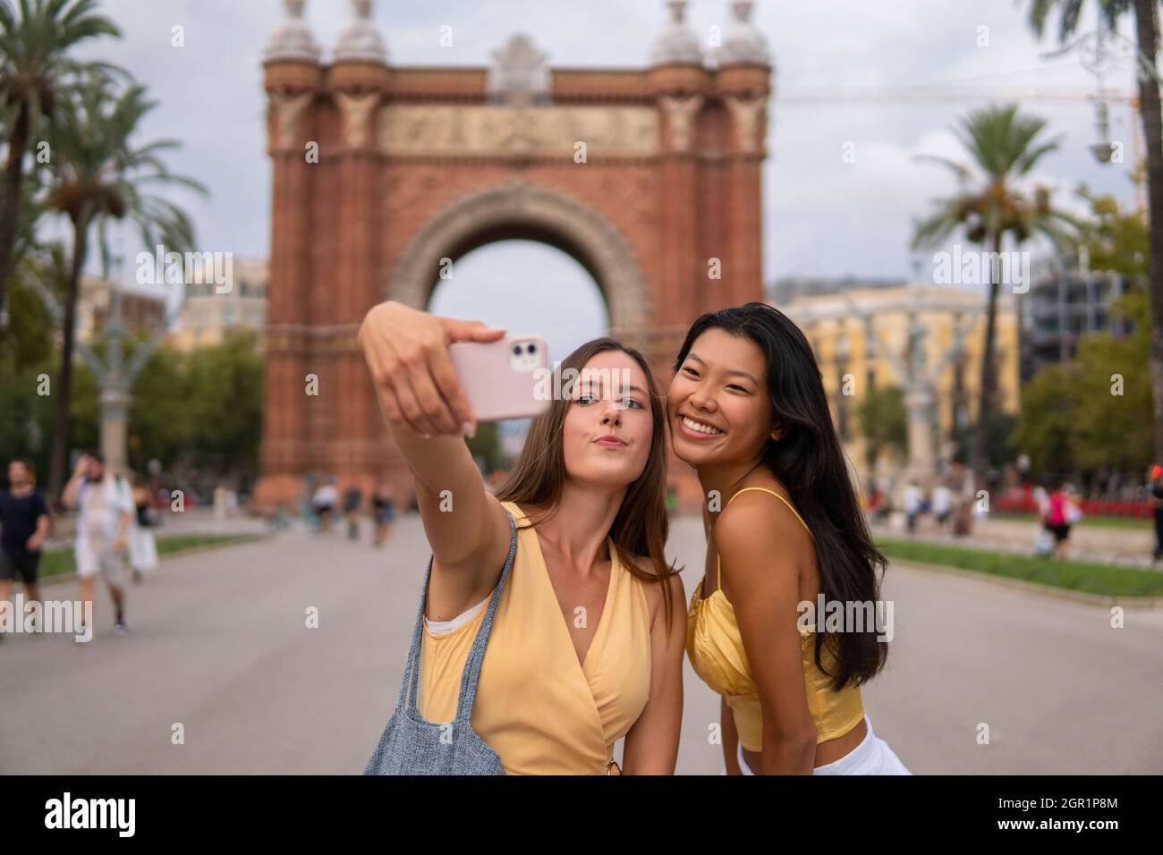 Cheerful young multiracial women in stylish clothes standing against triumphal arch of Barcelona city and taking selfie on mobile phone while enjoying summer journey in Spain Stock Photo