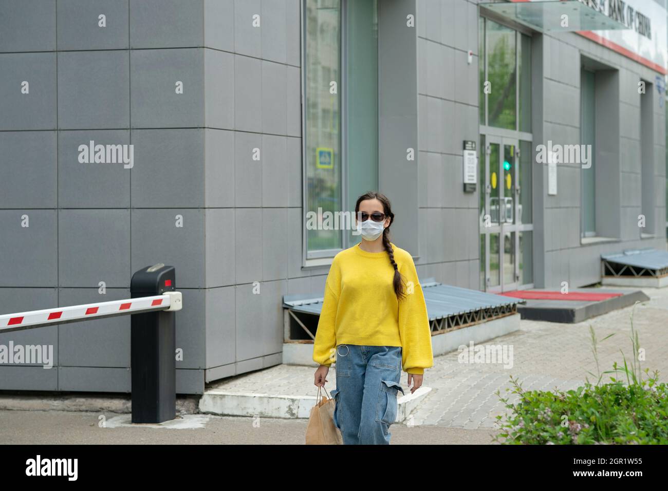 Girl In A Yellow Sweater And Jeans With A Mask And Sunglasses On Her Face Walks Past A Bank Of China Stock Photo