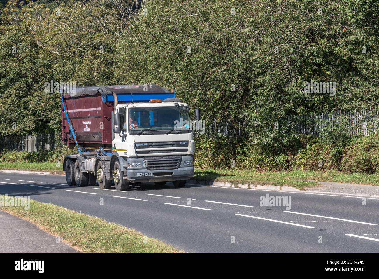 Front 3/4 view of unidentified DAF CF truck hauling a large industrial skip uphill on a country road. For UK construction, building trade activity. Stock Photo