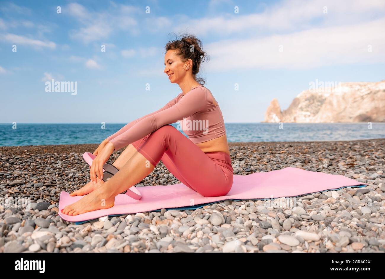 Young woman with black hair, fitness instructor in pink sports leggings and tops, doing pilates on yoga mat with magic pilates ring by the sea on the Stock Photo