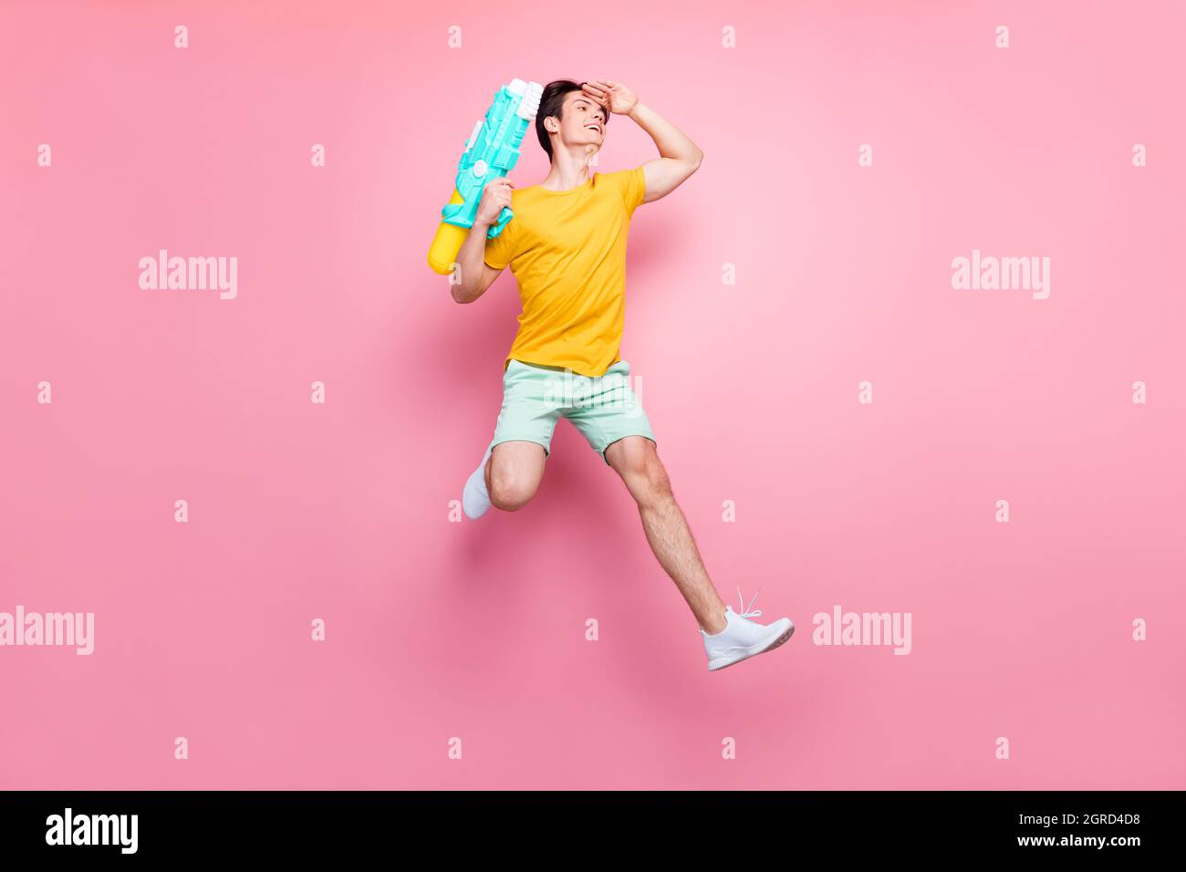 Full size photo of young guy happy positive smile jump look forward hold water pistol isolated over pink color background Stock Photo
