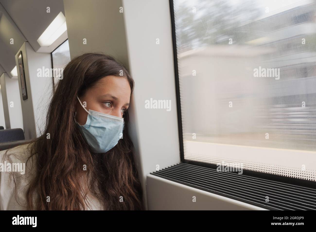 Pretty girl with long hair sitting in a local train looking outside the window. A young train passenger with protective mask. She seems to be travelin Stock Photo