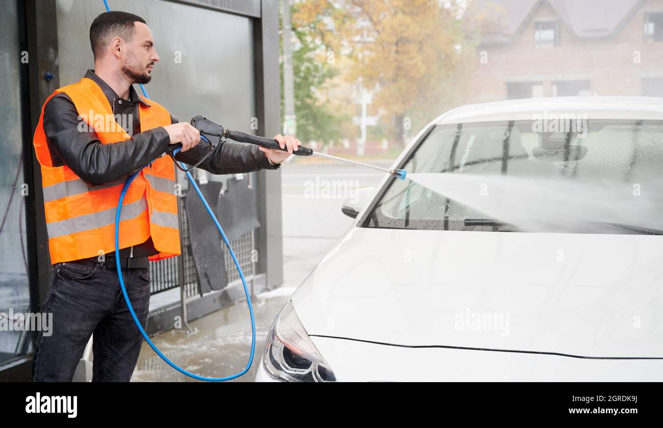 Young man washing white car on carwash station, wearing orange vest. Handsome worker cleaning automobile, using high pressure water. Stock Photo