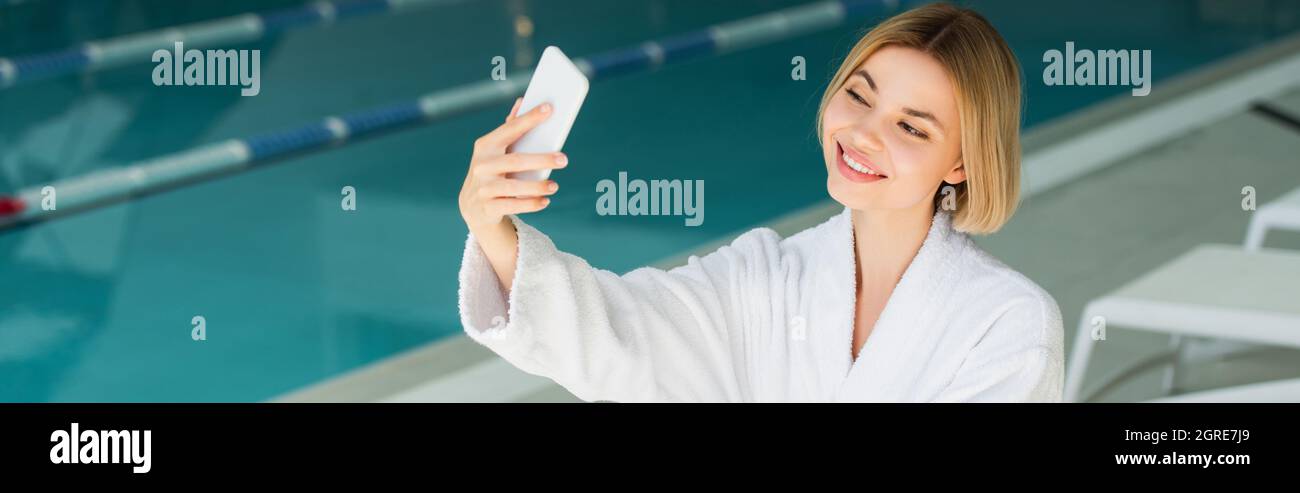 Cheerful woman in bathrobe taking selfie on smartphone near blurred