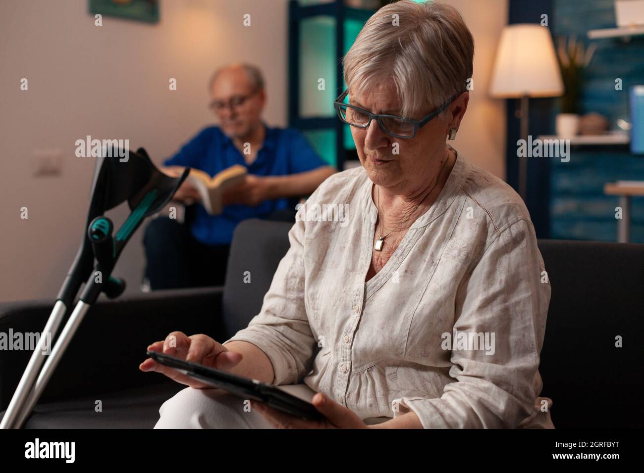Caucasian old woman holding modern tablet on sofa with crutches while elder disabled man sitting in wheelchair with book in living room. Senior married couple enjoying retirement at home Stock Photo