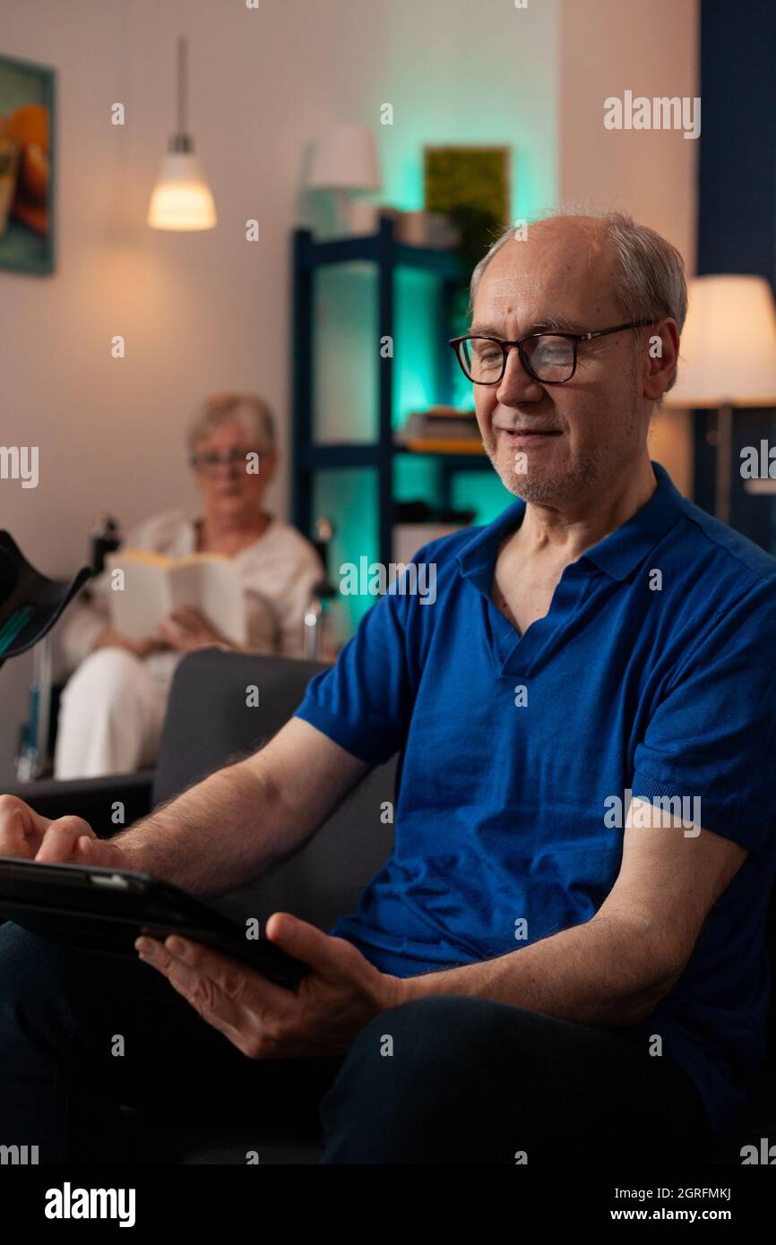 Close up of elder man holding and looking at digital tablet on living room sofa. Retired person with modern gadget at home and senior woman in background sitting in wheelchair. Stock Photo