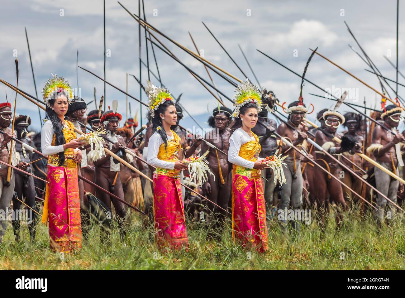 Indonesia, Papua, city of Wamena, Balinese women in traditional dress bringing a touch of national culture during a tribal war scene reenactment, by armed men from the Dani tribe. Baliem Valley Cultural Festival, every August, tribes come together to perform ancestral war scenes, parade and dance in traditional clothes Stock Photo