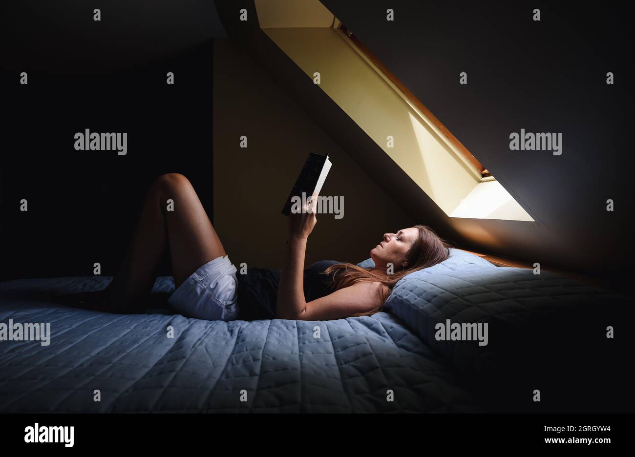 Woman laying on bed reading a book under a window in a dark room. Stock Photo