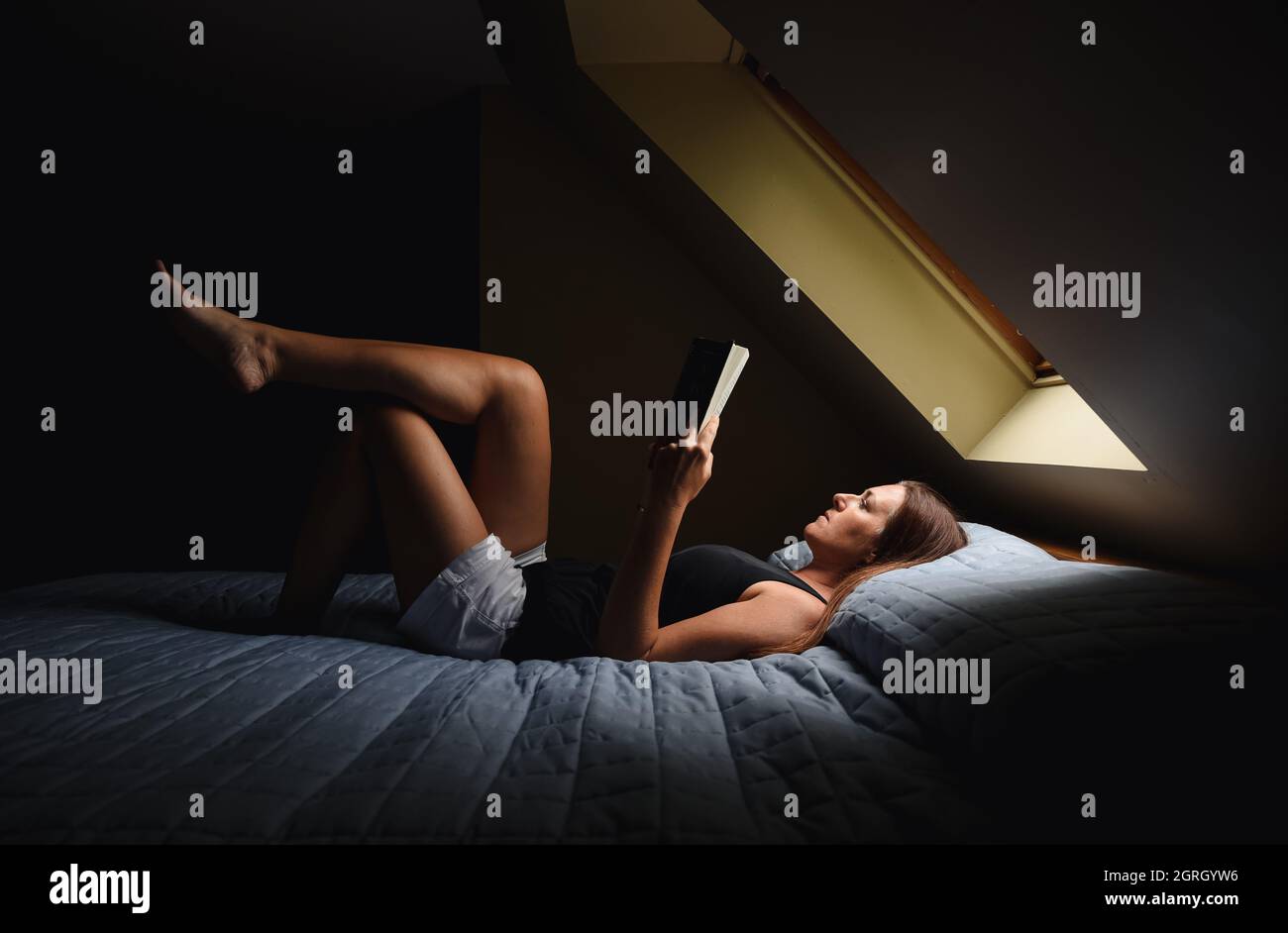 Woman laying on bed reading a book under a window in a dark room. Stock Photo