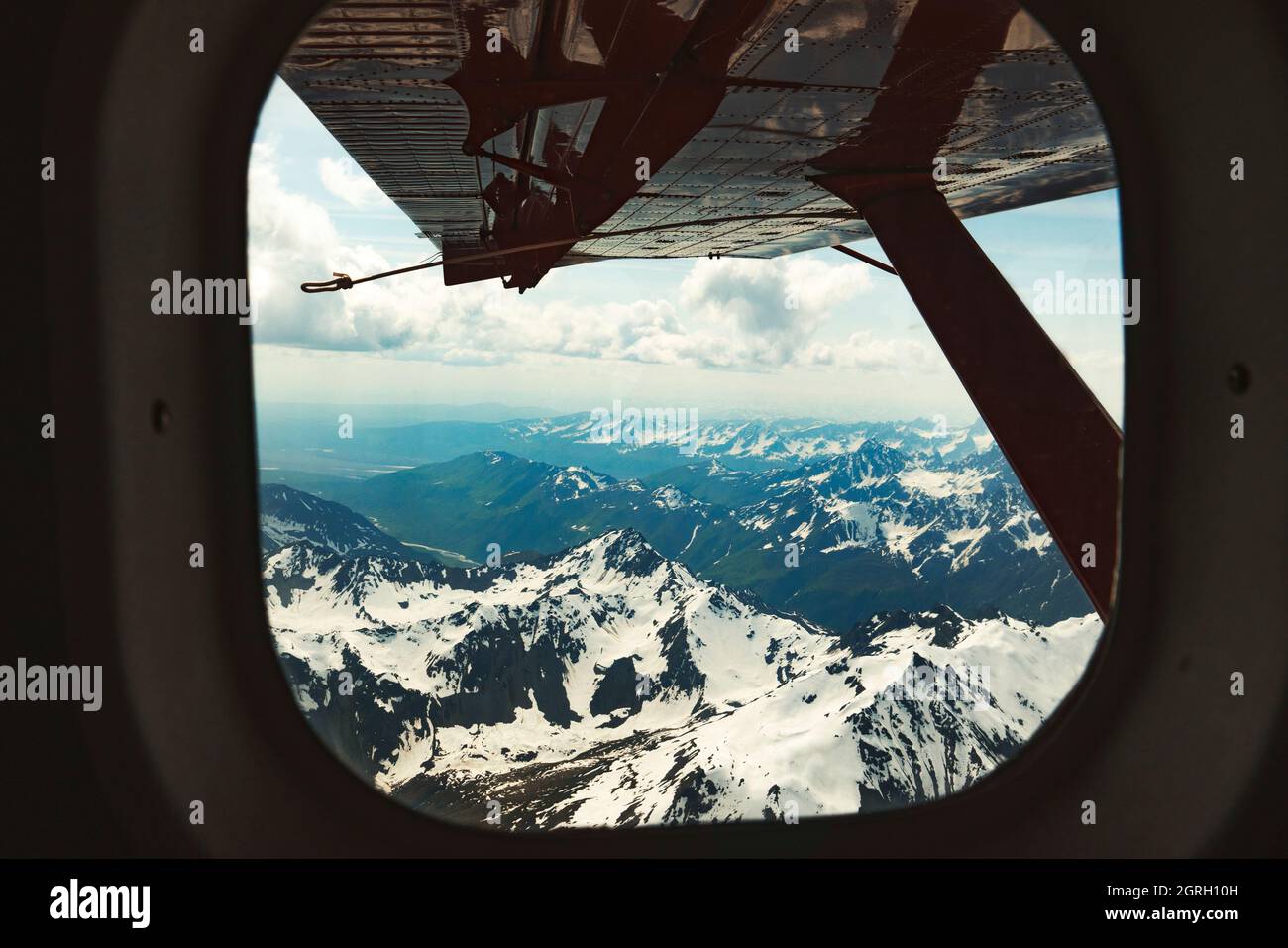 An airplane wing is seen out the window of a small plane with mountains in the distance Stock Photo
