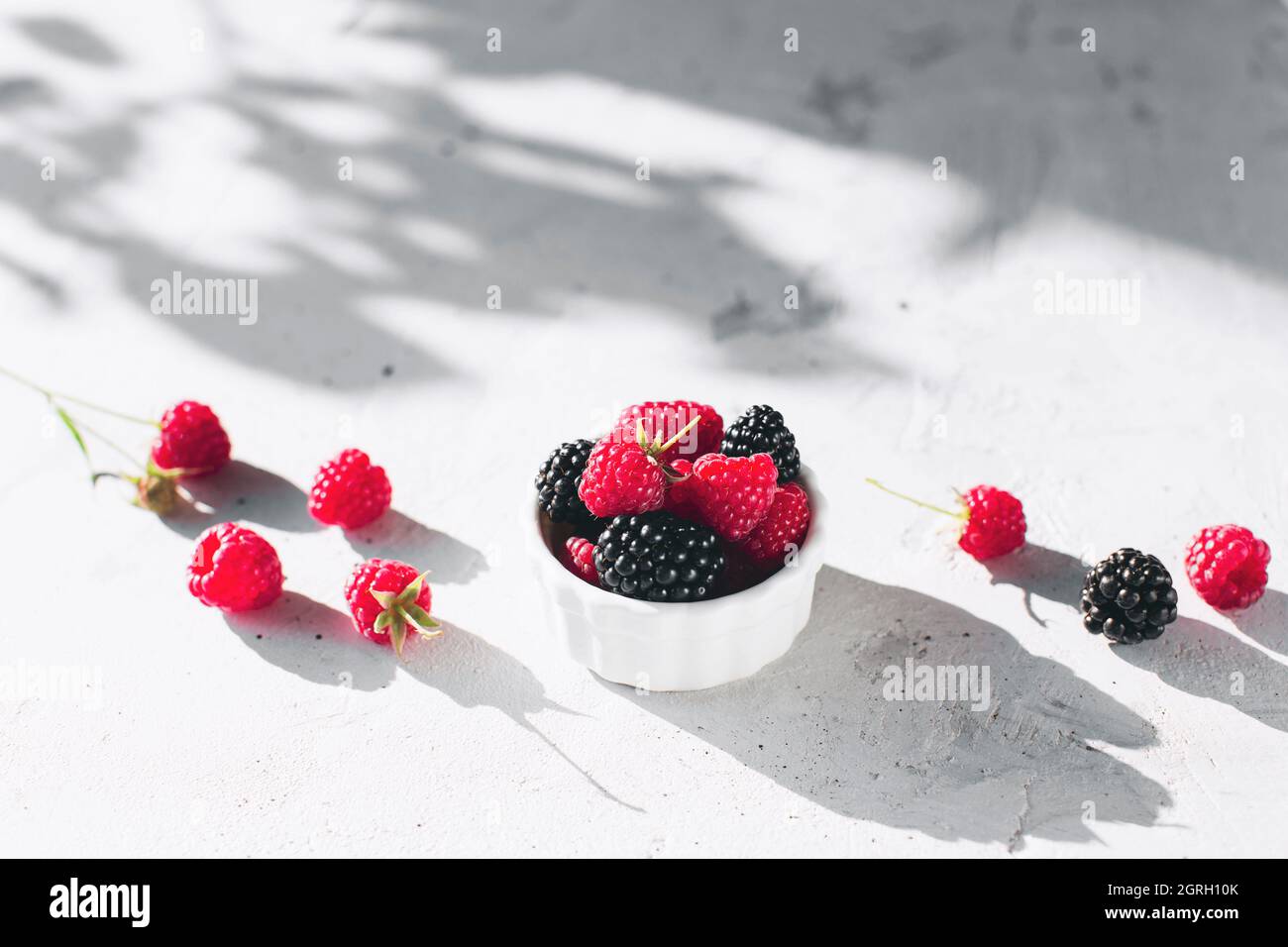 Bowl with raspberries and blackberries on gray concrete table with leaves shadow Stock Photo