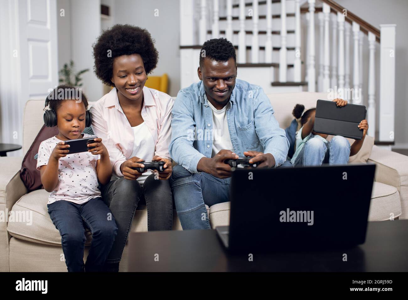 African wife and husband sitting on couch and playing video games on laptop  with joystick. Two cute daughters sitting near with tablet and smartphone  in hands. Modern technology for lifestyles Stock Photo -