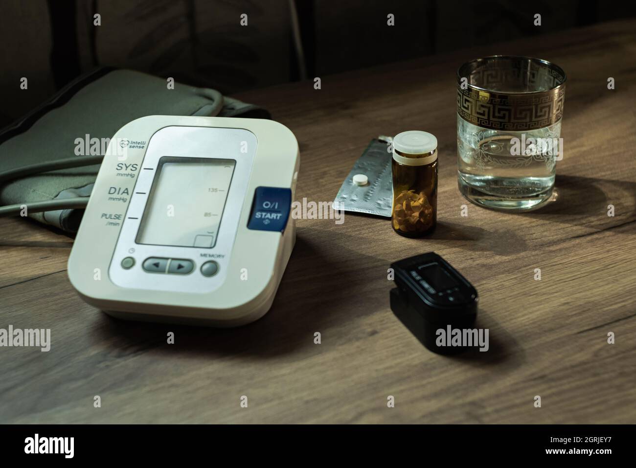 On a wooden table there is a blood pressure monitor, a glass of water, pills, and an oxyometer. Health Concept Stock Photo
