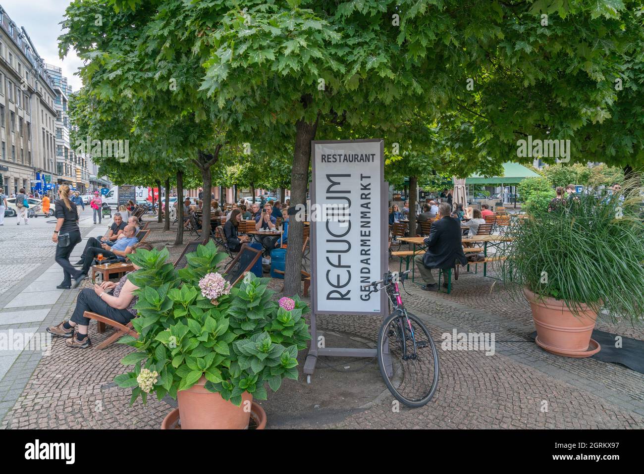 Berlin Germany - August 26 2017; People seated relaxing under trees along street and in Gendarmenmarkt Square by sign for Refugium Restaurant. Stock Photo