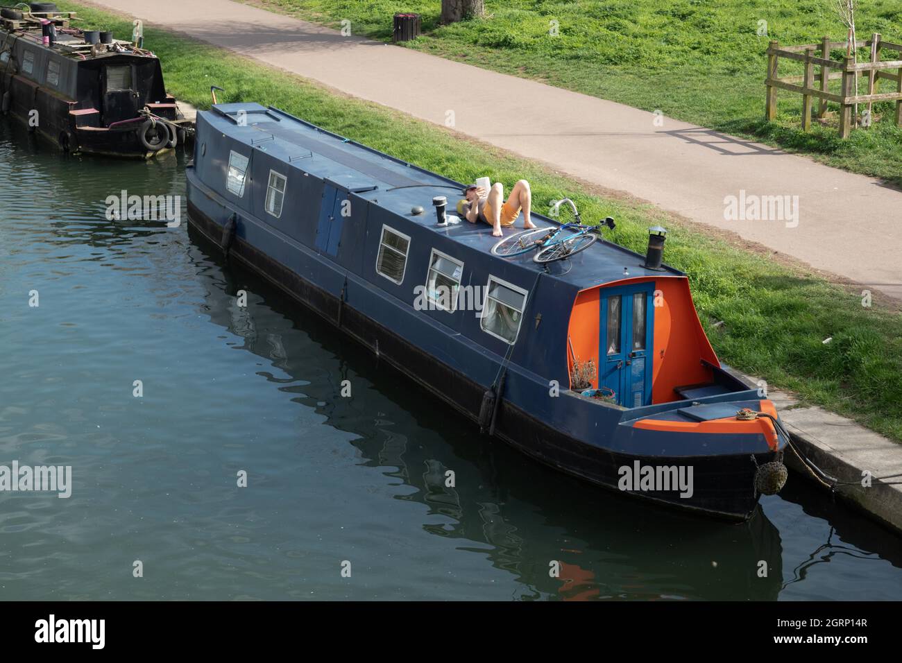 Blue narrowboat moored on River Cam with a man lying on roof relaxing reading with bicycle also on roof Cambridge England Stock Photo