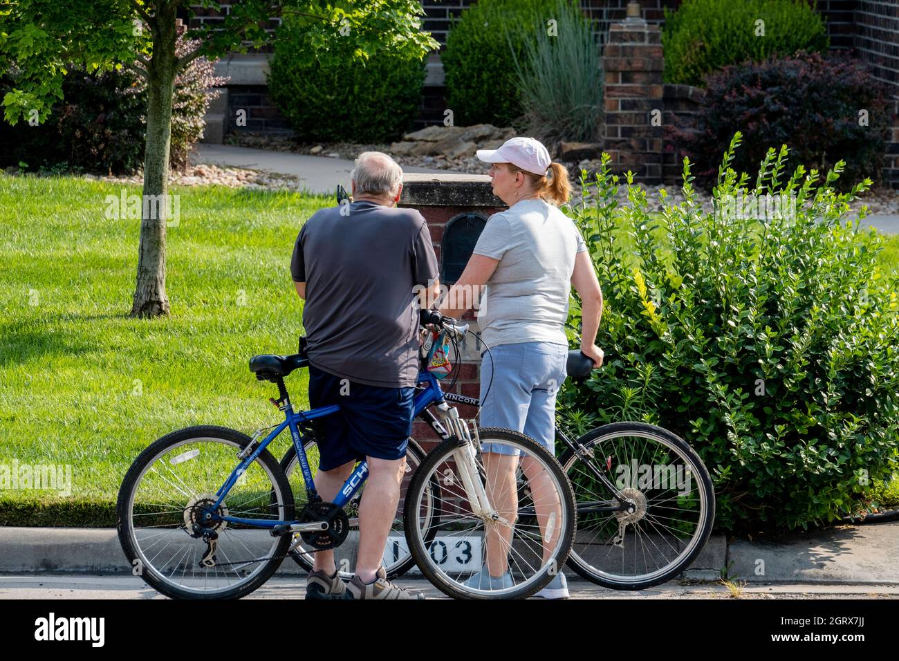 A senior Caucasian man stops his bicycle in the street of a residential neighborhood to answer his mobile phone while a woman companion waits. USA. Stock Photo