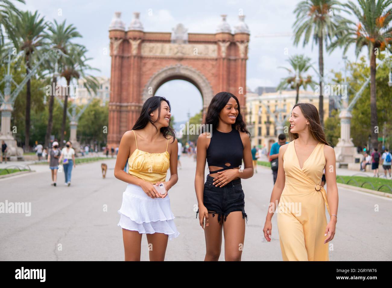 Young multiracial girlfriends in stylish fashionable clothes talking and laughing while strolling on square of Barcelona city with triumphal arch in background in summer day Stock Photo