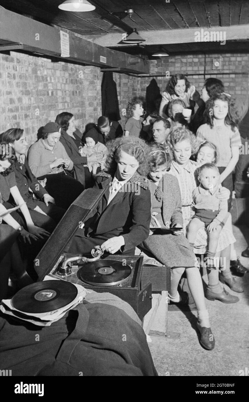 A young woman plays a gramophone in an air raid shelter in north London during 1940. A young woman places the gramophone needle on a record to bring some light relief to an air raid shelter, somewhere in north London. The rest of the shelterers appear to be enjoying her choice of music. In the background, one woman can be seen knitting, as others chat to pass the time. Stock Photo