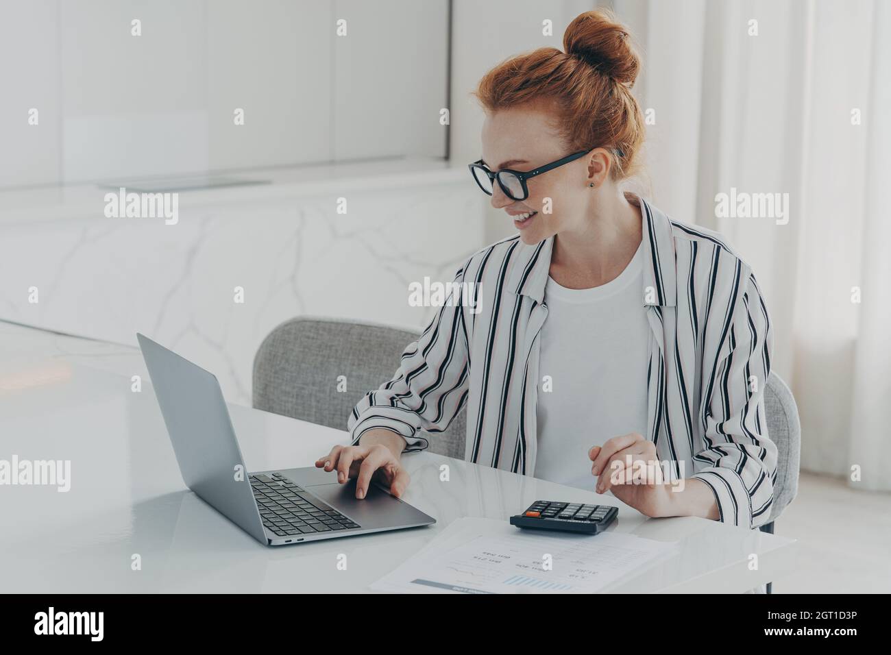 Young positive red-haired woman accounter working remotely on laptop and using calculator Stock Photo