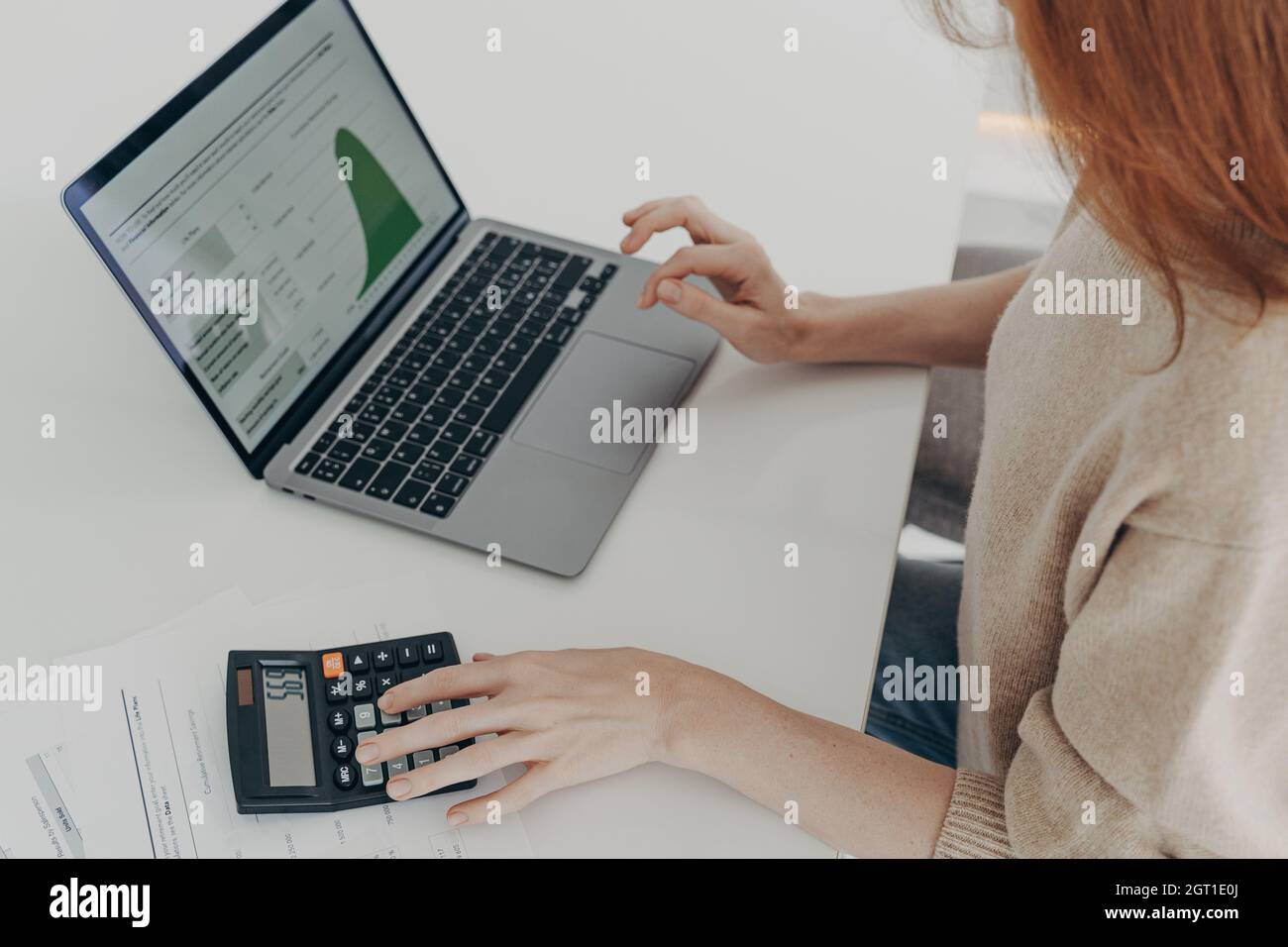 Woman using laptop and calculator while sitting at table at home, looking at screen with diagrams Stock Photo