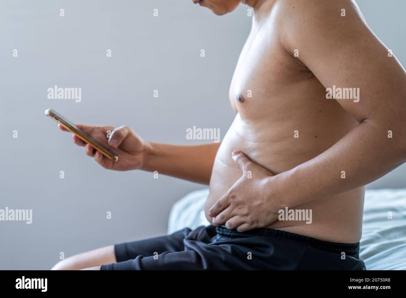 Big Belly Man Sitting On Bed Using Smart Phone. Stock Photo