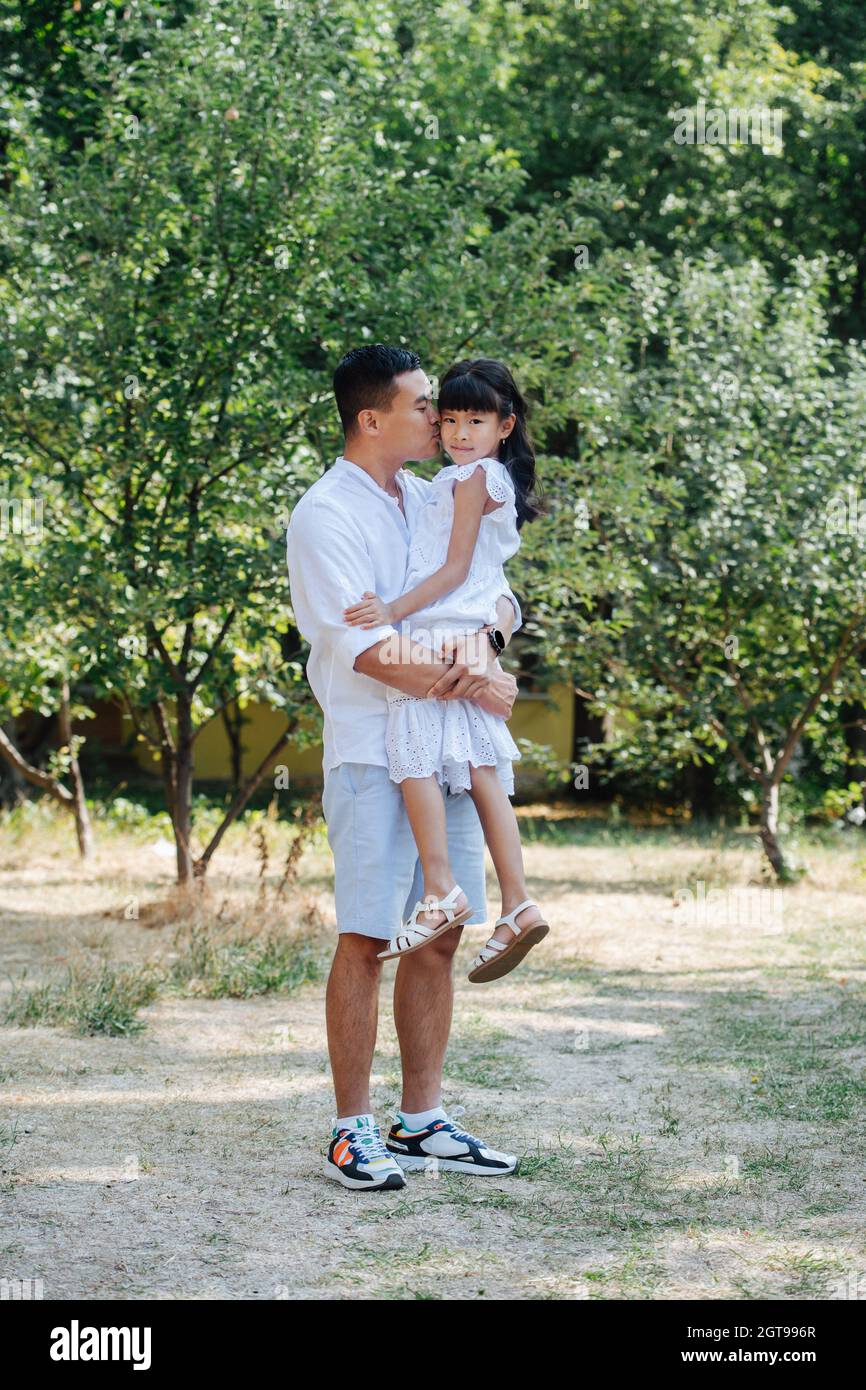 Longshot of an asian father holding his daughter in his hands, kissing her in a cheek. She's looking at the camera. They are spending time in a park, Stock Photo