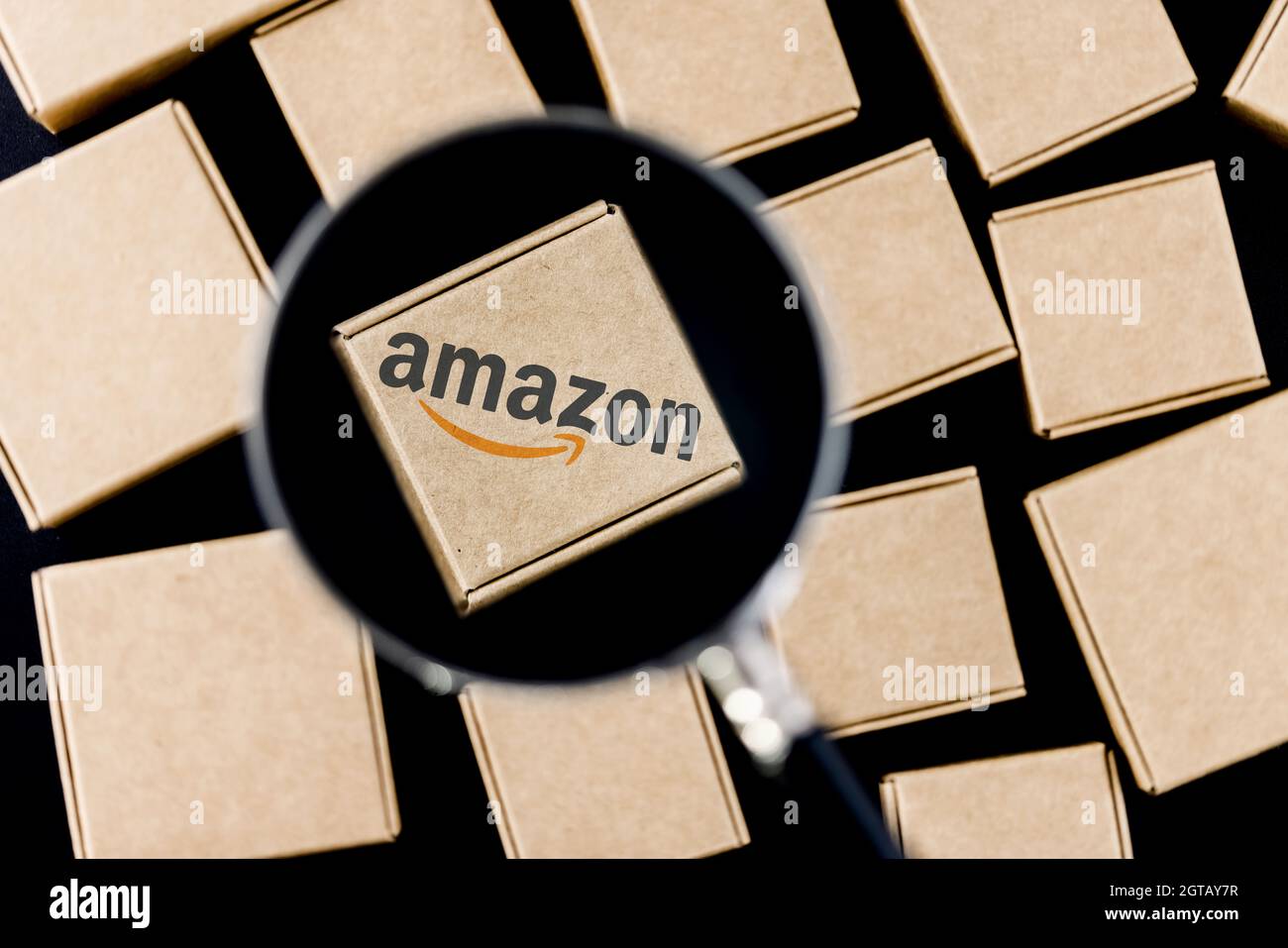 Modified photo of Amazon logo on a box under a magnifying glass surrounded by parcels. Stock Photo