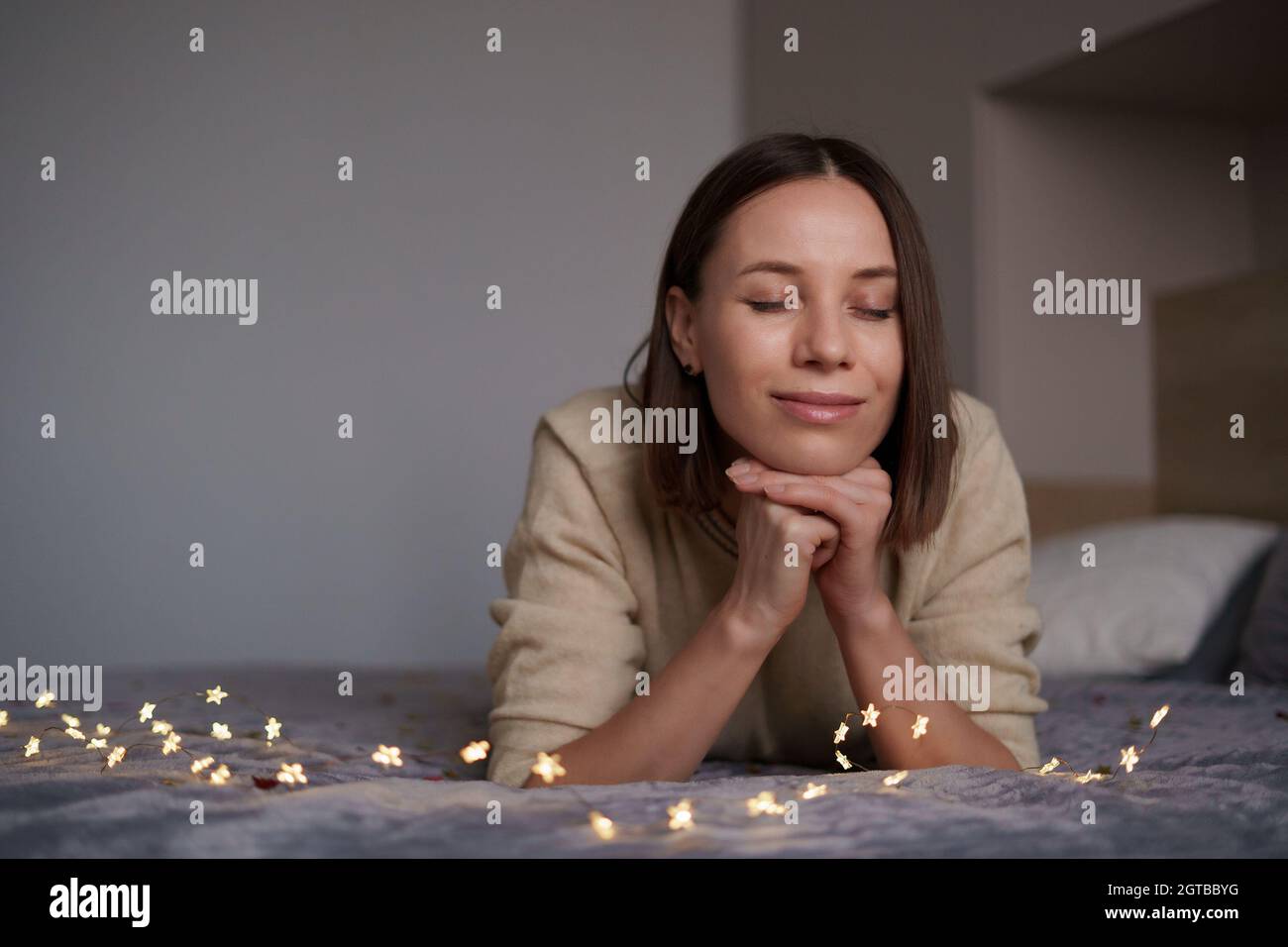 Pretty Caucasian woman smiling with fairy lights laying on bed. Comfy home. Dreaming time Stock Photo