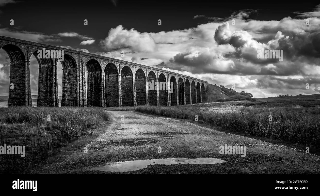 Black and white image of the Ribblehead Viaduct Yorkshire UK. with open road and water pools. Clear skies but dramatic clouds. No people. Stock Photo