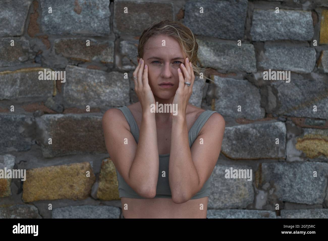 Sad stressful face of young caucasian blonde woman in crop top on the stone background. Young woman crying and using hands to cover face. Stock Photo
