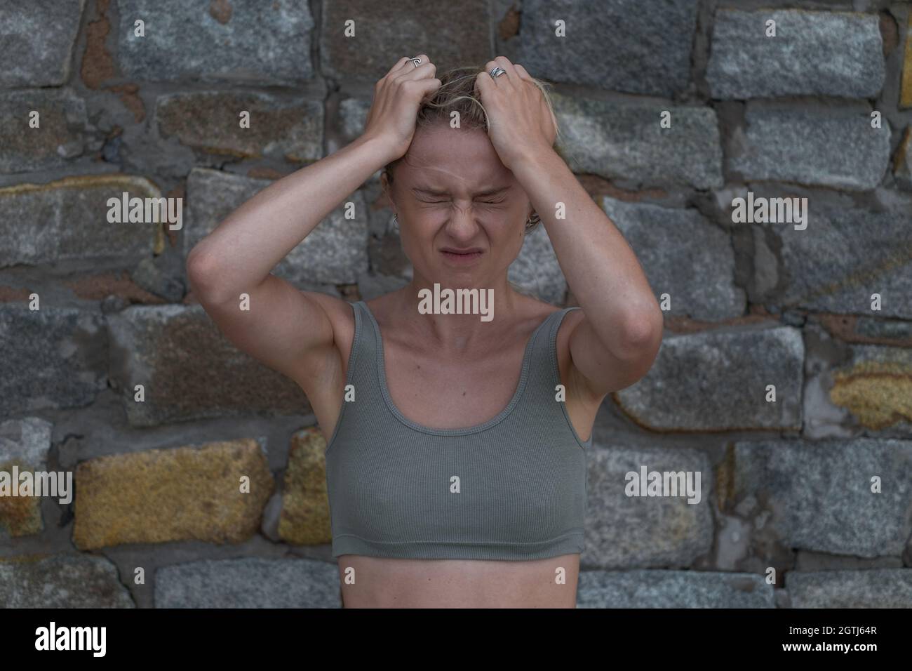 Portrait of stressful angry young caucasian blonde woman in crop top on the stone background. Angry girl tears the hair on her head. panic attacks  Stock Photo